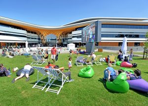 Image shows exterior of Bunjil Place Plaza with grass and people relaxing in deck chairs and beanbags in the sunshine