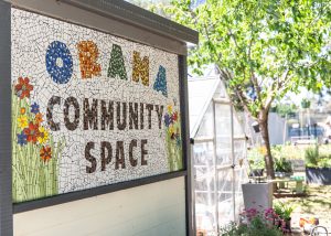 Mosiac tiled signage with words Orana Community Space and a greenhouse and gardens in the background