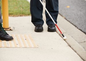 Person using a white cane standing next to tactile ground surface indicators at a road crossing