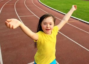 Young girl with Down Syndrome wearing bright yellow t-shirt and running on an athletics track with arms in the air and an happy expression on her face