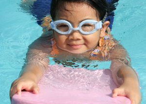 Young girl swimming in the water wearing swimwear and goggles and holding a kickboard