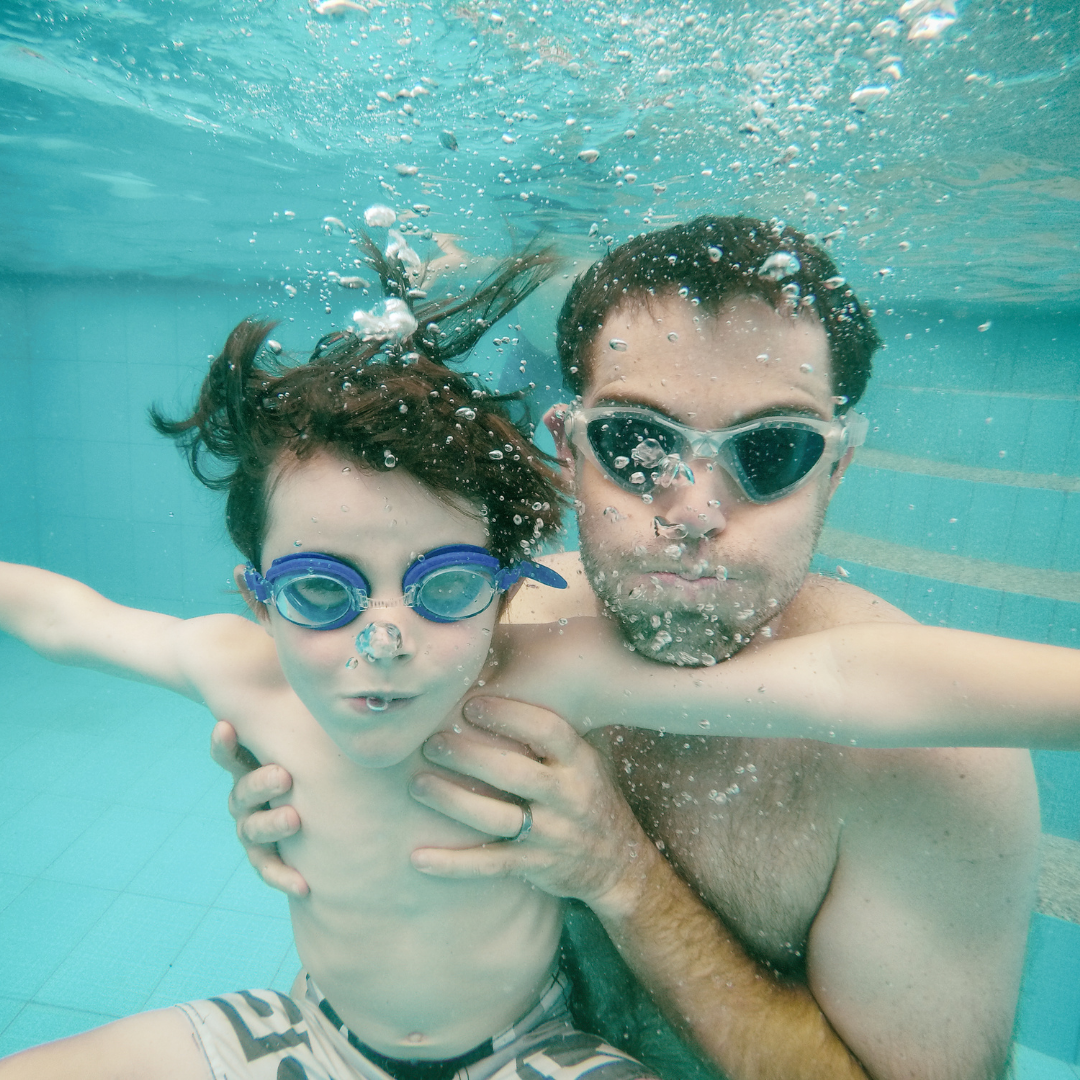 Father and son wearing bathers and goggles swinning underwater in a pool and blowing bubbles