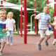 three young children running at a playground with play red play equipment in the background