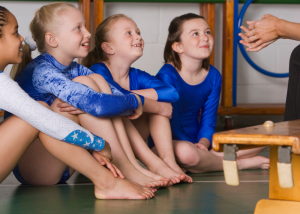 Four young girls wearing blue leotards, sitting on floor in gymnastics studio listening to a coach who is sitting on a bench