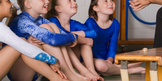 Four young girls wearing blue leotards, sitting on floor in gymnastics studio listening to a coach who is sitting on a bench