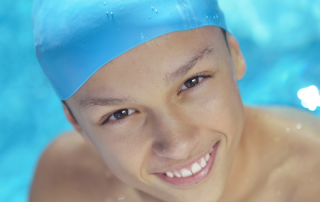 Young boy wearing a pale blue swimming cap pictured in the water and smiling at camera