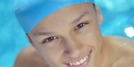 Young boy wearing a pale blue swimming cap pictured in the water and smiling at camera