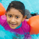 Young asian girl wearing colourful bathers and arm bands swimming in a pool looking at camera and smiling
