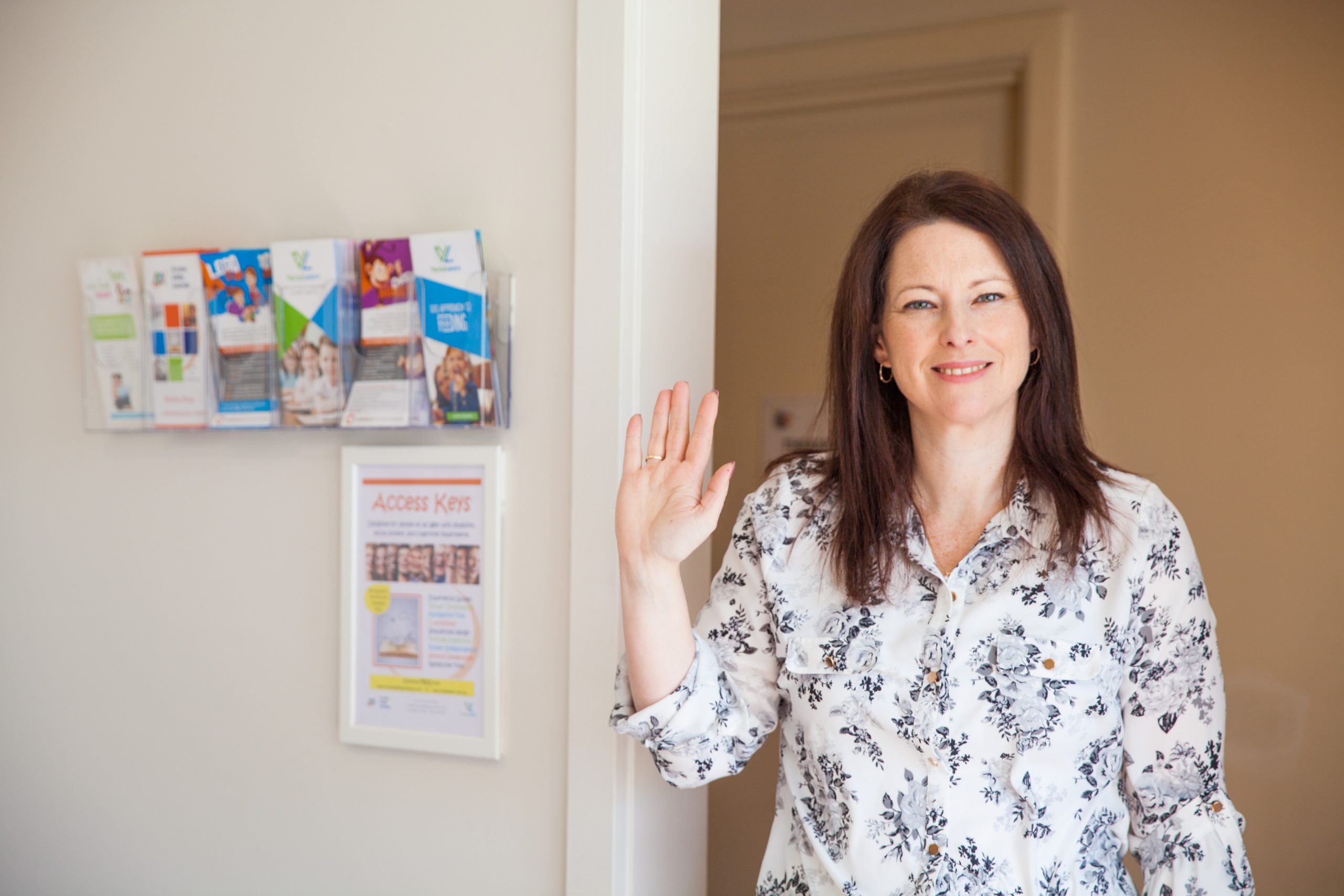 Female speech pathologist standing in speech pathology clinic waving. Background with Access Key signage and brochures