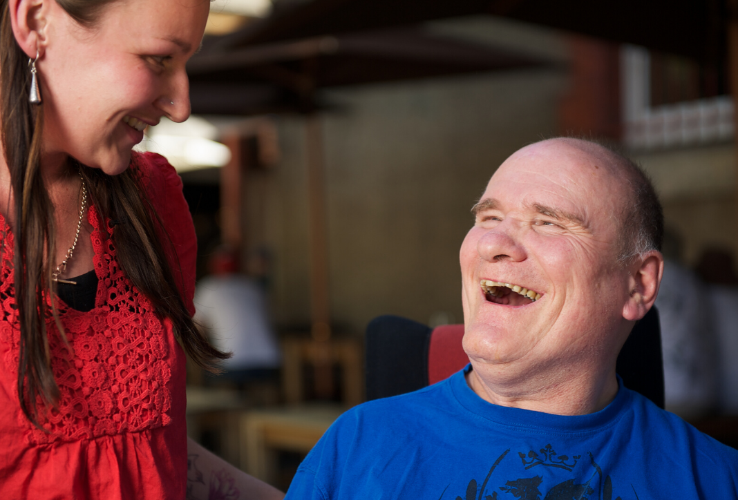 Man smiling wearing blue t-shirt seated in wheelchair. Female disability support worker standing next to him wearing a red shirt