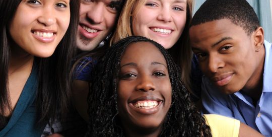 Five young adults huddled in a group facing the camera and smiling