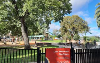Lake Weeroona Play Space gate with signage and surrounding play space fencing with trees and play equipment in the background