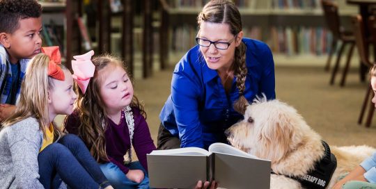 A librarian in a library reading a story to a group of smalle children. Seated next to her is a therapy dog