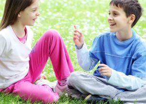 Two children, a girl and a boy, sitting on grass engaging in coversation and both smiling