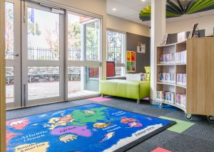 Interior or Broadford Library with bookshelves, books, rug on floor and glass windows and doors leading to outdoor area