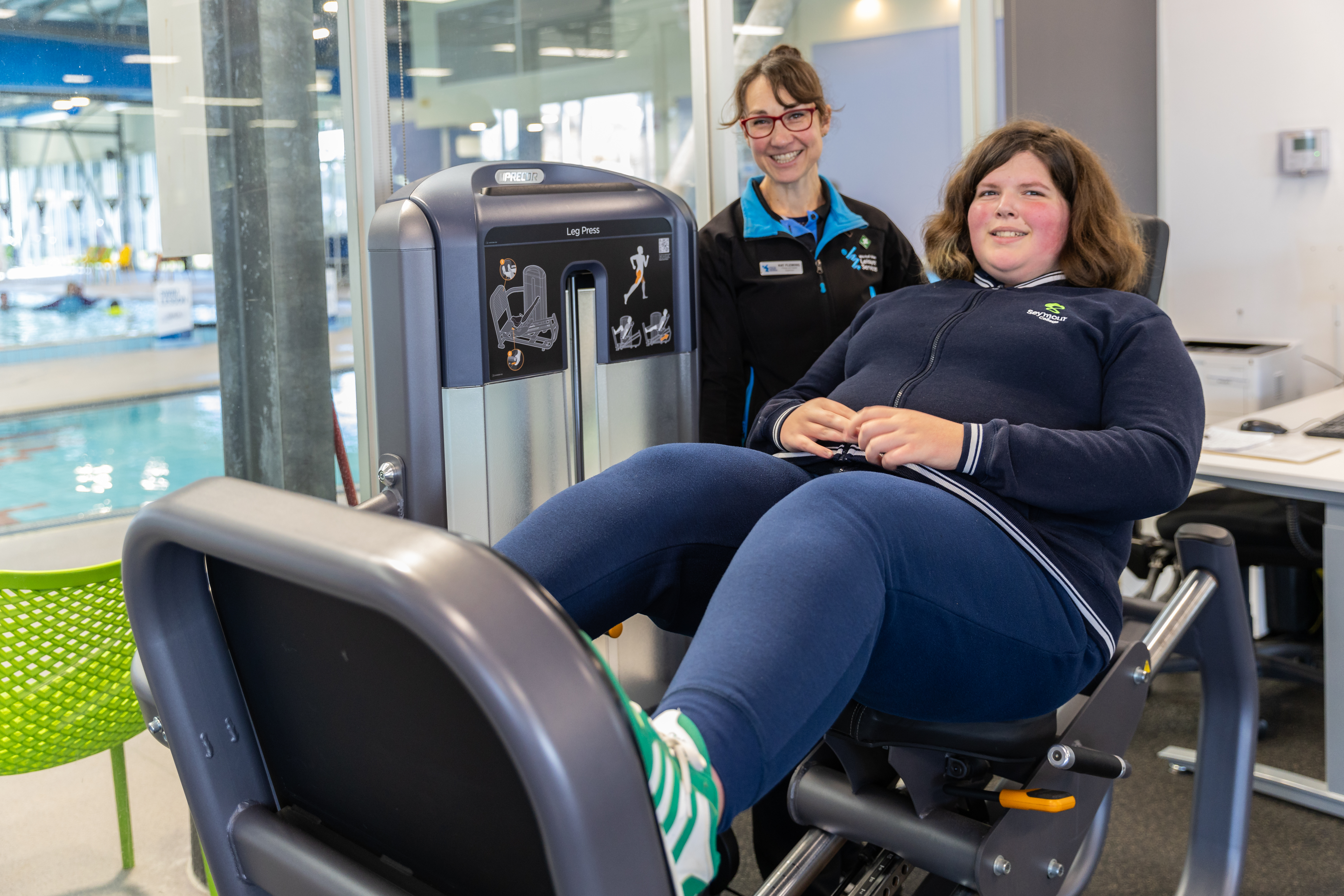 Young female student on gym equipment with personal trainer in background